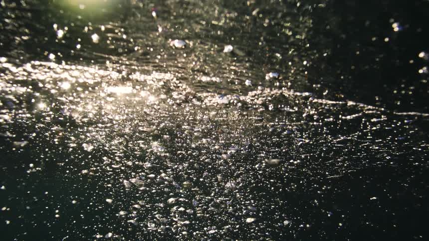 An abstract slow-motion view from underwater as bubbles rise towards the bright sun, with the camera eventually breaking the surface