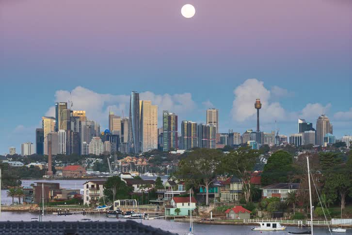 A day-to-night timelapse of the Sydney city skyline with a full moon rising during a colorful sunset over the harbor