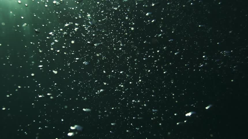 An abstract slow-motion shot of countless air bubbles rising through dark, sunlit water