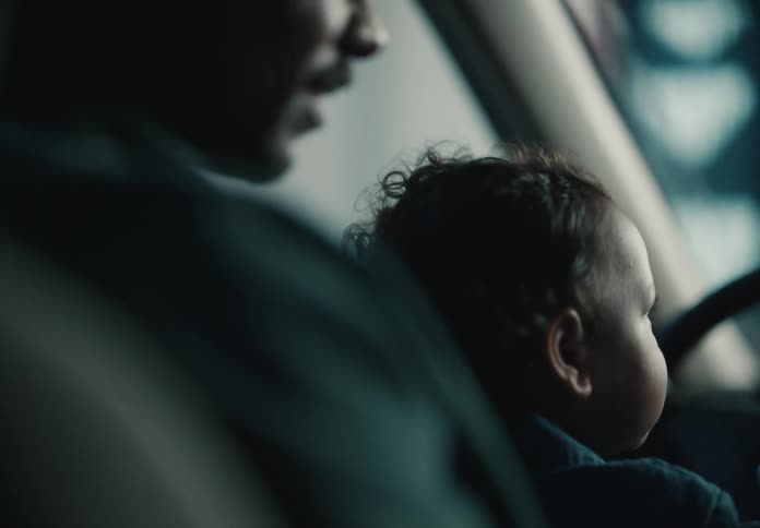 A father holds his baby with curly hair in the driver's seat of a car in a dimly lit setting