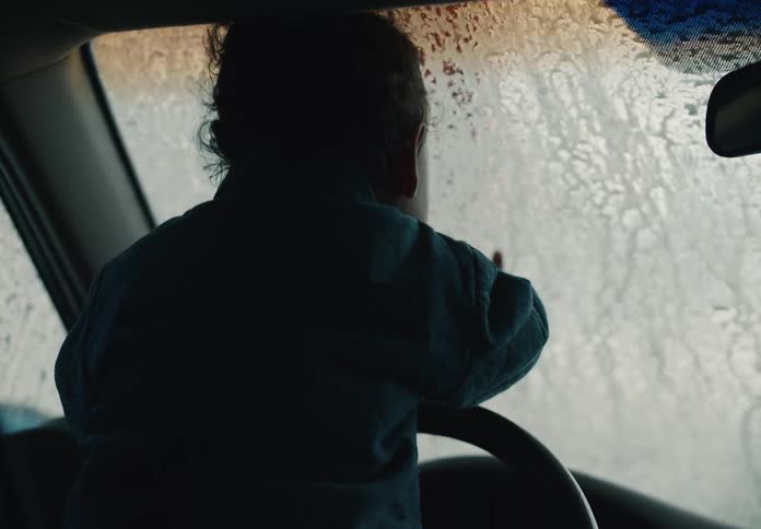 A curious toddler sitting in a car's driver seat watches and touches the sudsy windshield during a car wash