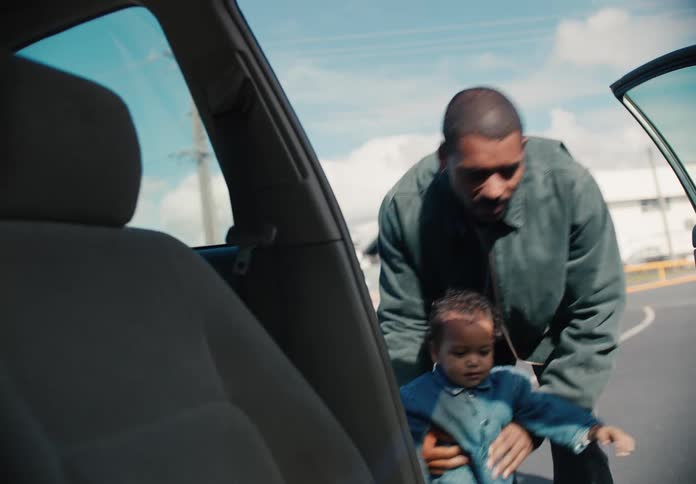 A father holds his toddler's hand and walks towards an open car door before lifting the child inside