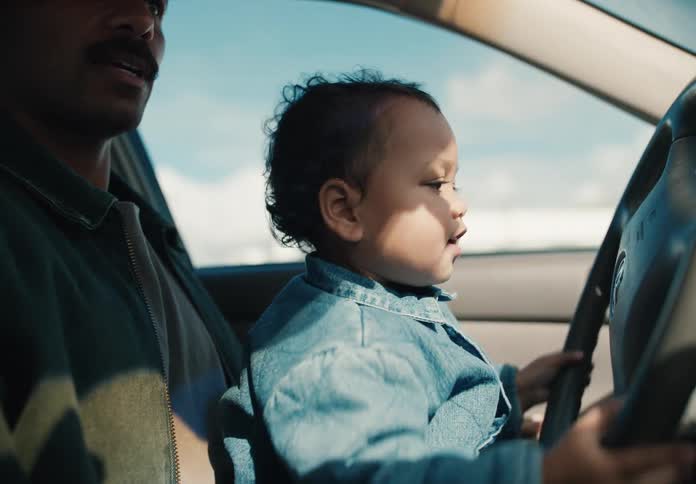 A happy toddler sits on his father's lap in the driver's seat, playfully pretending to steer the car on a sunny day