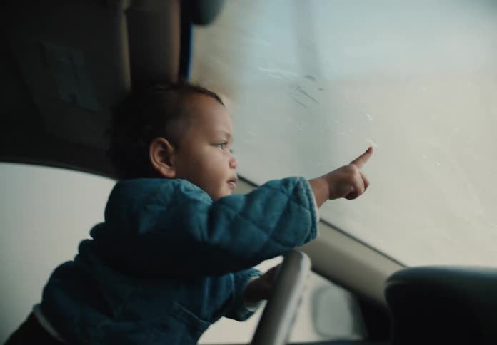 A father holds his curious toddler on his lap as the child pretends to steer the car, pointing out the window