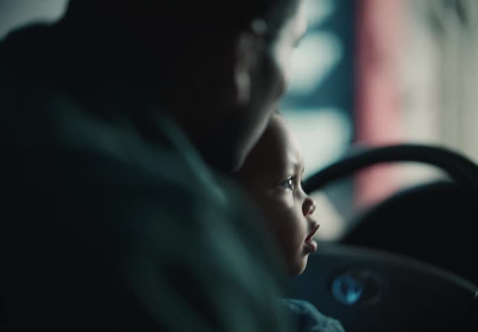 A curious baby sits on their parent's lap, gripping the steering wheel while pretending to drive the car