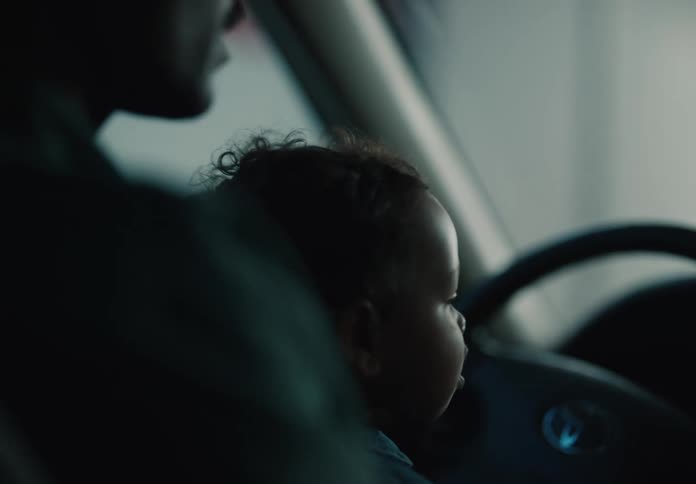 A caring father holds his baby on his lap while sitting in the driver's seat of their car in a dimly lit setting