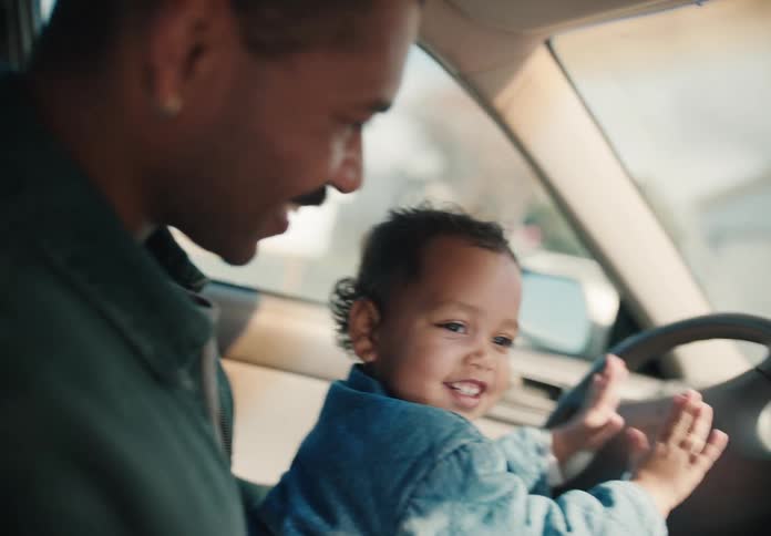A happy toddler smiles as he sits on his father's lap in the driver's seat, pretending to drive the car