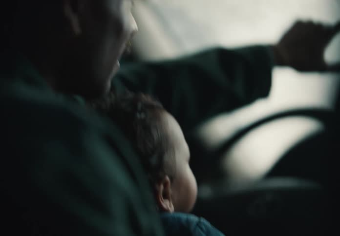 A father holds his baby on his lap, teaching them to drive as they sit together in the front seat of a car