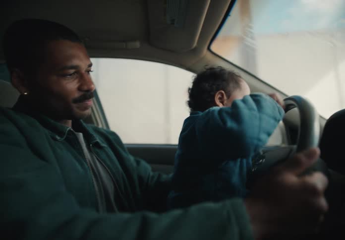 A happy father watches as his toddler sits on his lap and pretends to drive the car