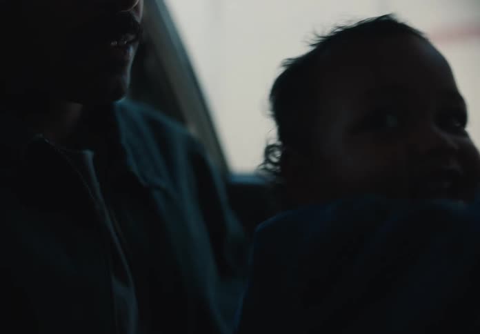 A young child sits on their father's lap in a dimly lit car, curiously watching the world go by outside the window