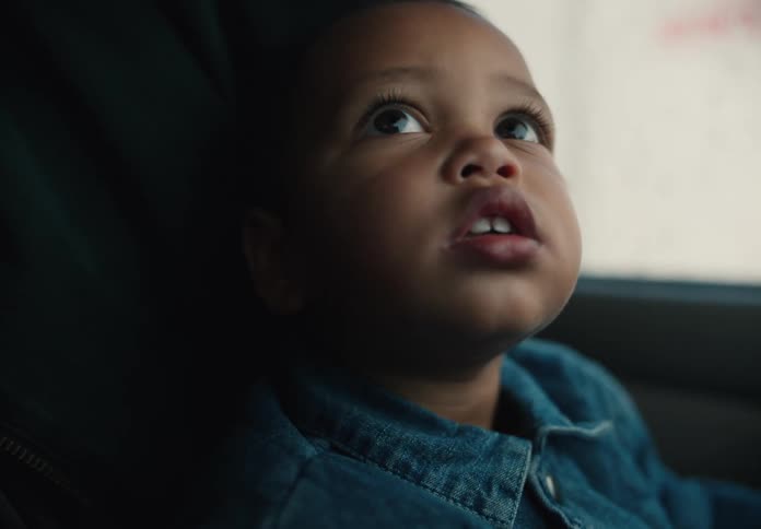 An adorable toddler in a denim jacket gazes up with wonder while sitting on his father's lap in a car