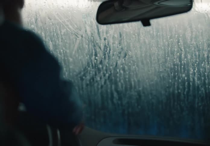 A father holds his young child on his lap as they both point at and draw on the soapy windshield while going through a car wash