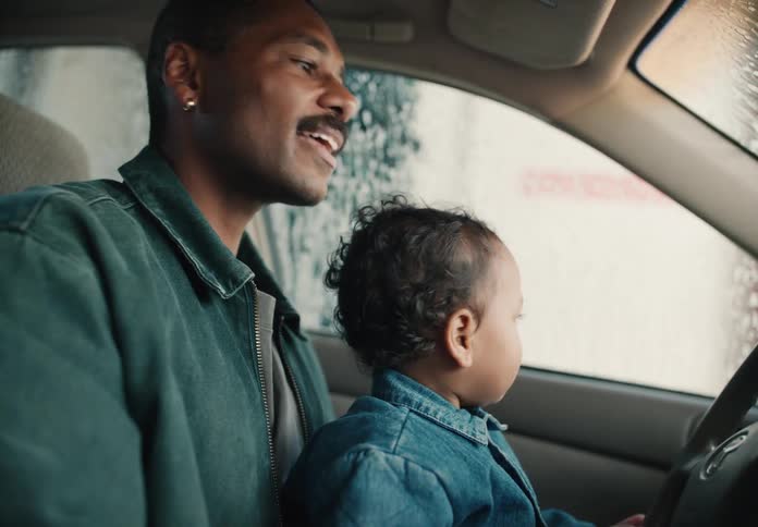 A smiling father holds his curious toddler on his lap as they watch the water and soap run down the windows in an automatic car wash