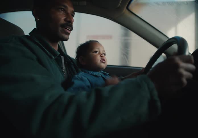 A smiling Black father holds his curious toddler on his lap, letting the child pretend to drive as they go through a car wash