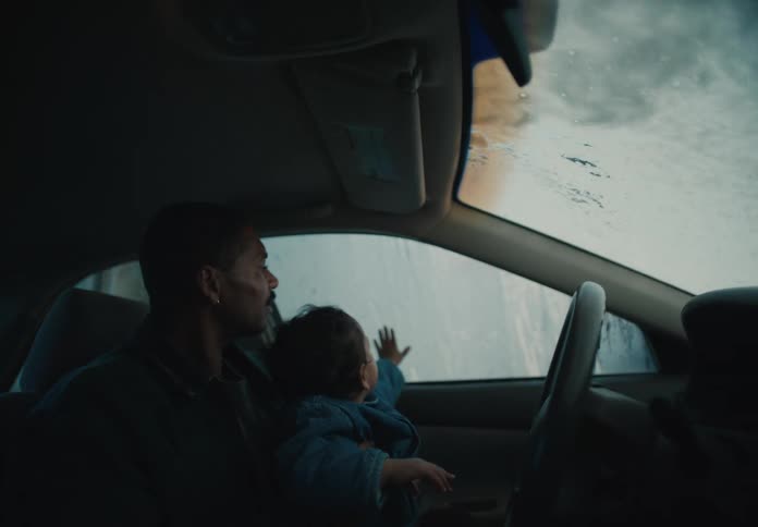 A curious toddler sits on their father's lap, mesmerized by the sights and sounds of an automatic car wash from inside the car