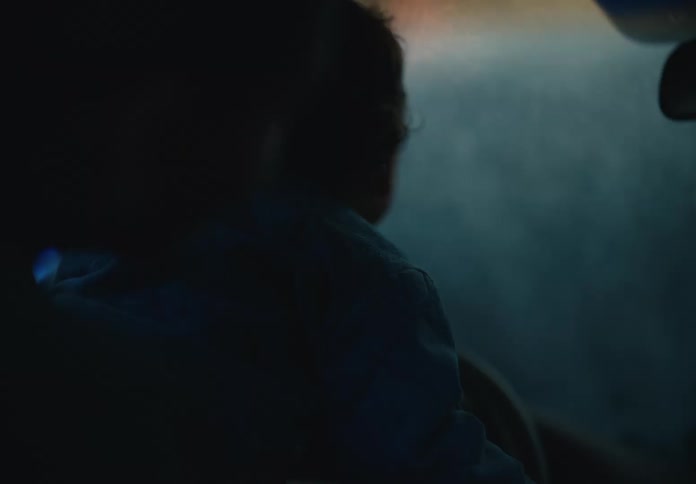 A slow-motion, over-the-shoulder shot shows a parent and child in the driver's seat of a car during a dark and moody night drive