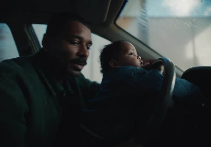 A loving father holds his baby on his lap in the driver's seat, pointing out the window and talking to them