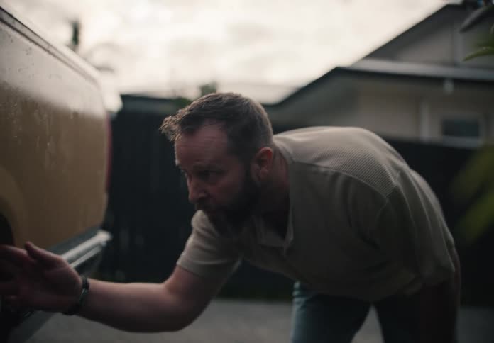 A man with a beard inspects the rear of his yellow pickup truck in the driveway as it starts to rain