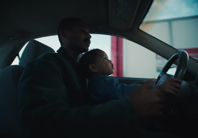 An African American father holds his curious young child on his lap as they drive through an automatic car wash, sharing a special bonding moment