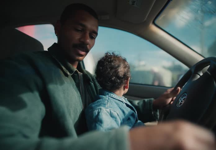 A young father drives his car while his adorable toddler sits on his lap, sharing a heartwarming moment together