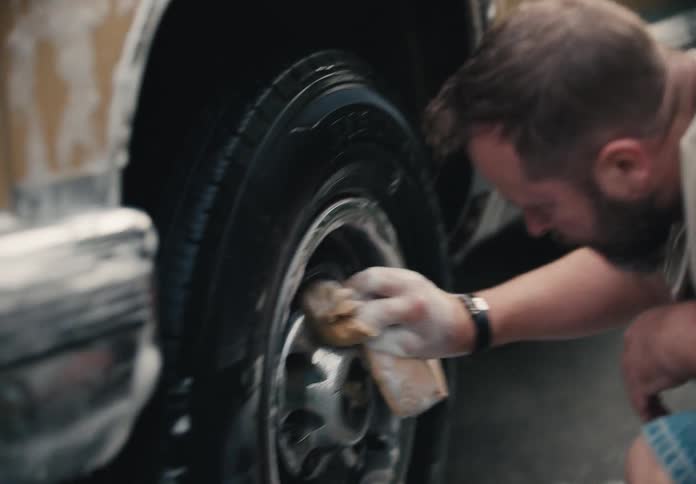 A bearded man kneels down and scrubs the soapy wheel of his truck with a sponge