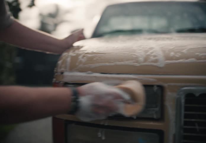 A man diligently washes the soapy hood and headlights of his classic tan GMC truck in the driveway