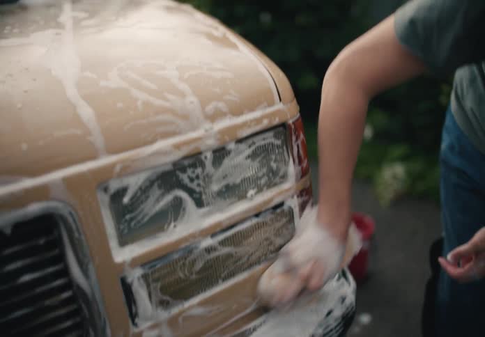 A detailed slow-motion close-up of two people scrubbing the soapy headlights of an old tan truck