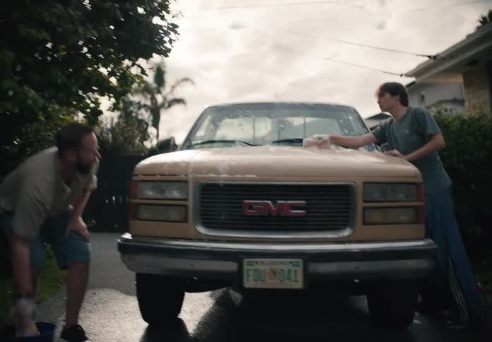 A father and his teenage son wash their vintage tan GMC pickup truck together in the driveway of their suburban home