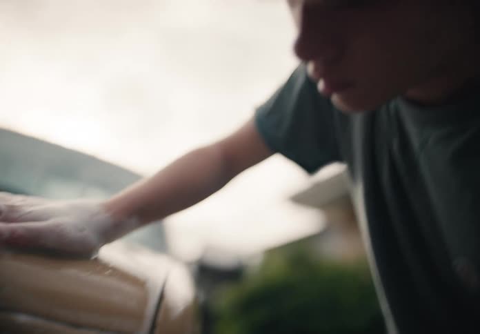 A close-up reveals a teenage boy diligently washing a vintage yellow car with soapy sponges