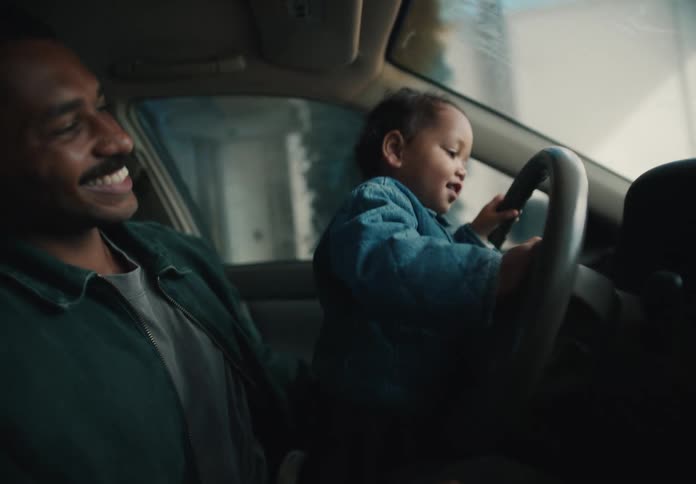 An adorable toddler joyfully pretends to drive while sitting on their father's lap inside a car
