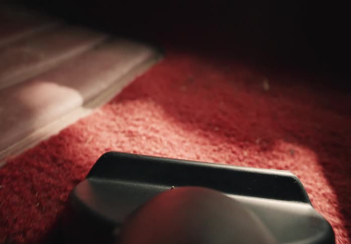 A first-person view from a vacuum cleaner as it cleans a red carpet in a sunlit room