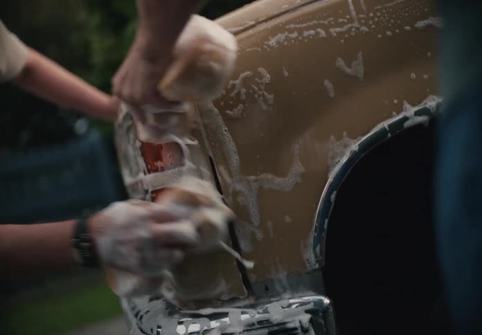 A bearded father and his son bond as they carefully wash the taillight of their classic car with soapy sponges in their backyard