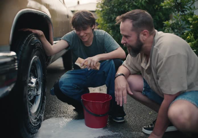 A father and his teenage son share a happy moment while washing the tires of their vintage truck in the driveway