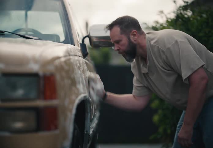 A bearded man in casual clothes washes his vintage pickup truck in the driveway with a soapy sponge in slow motion