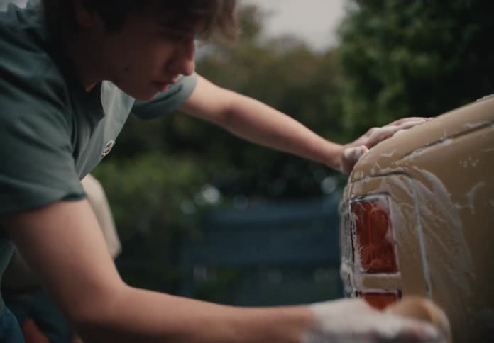 A teenage boy carefully washes a car with a soapy sponge in slow motion as his father helps in the background