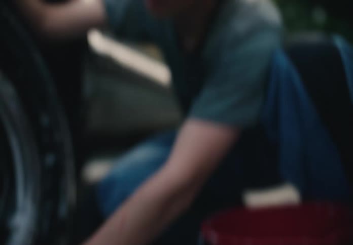 Rack focus from a hose nozzle on the pavement to a teenage boy diligently washing the tires of a car