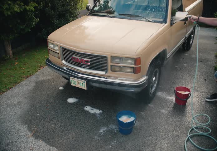 A man hoses down his vintage tan pickup truck in a suburban driveway