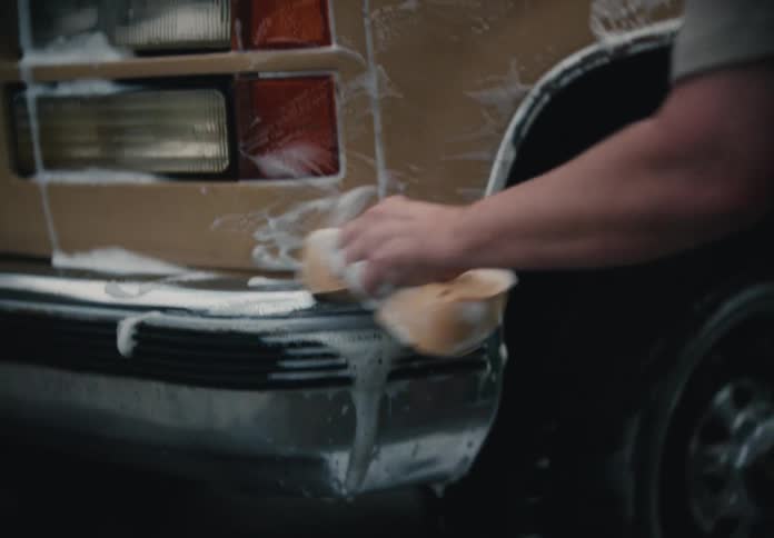 A man diligently scrubs the soapy front end of his vintage tan truck with a sponge