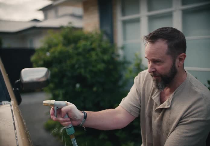 Bearded man in his driveway intently rinses the soap suds off his vintage truck in slow motion