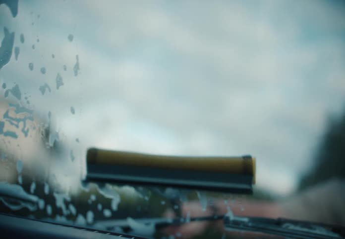 A squeegee slowly cleans a soapy car windshield in a cinematic slow-motion close-up shot from inside the vehicle