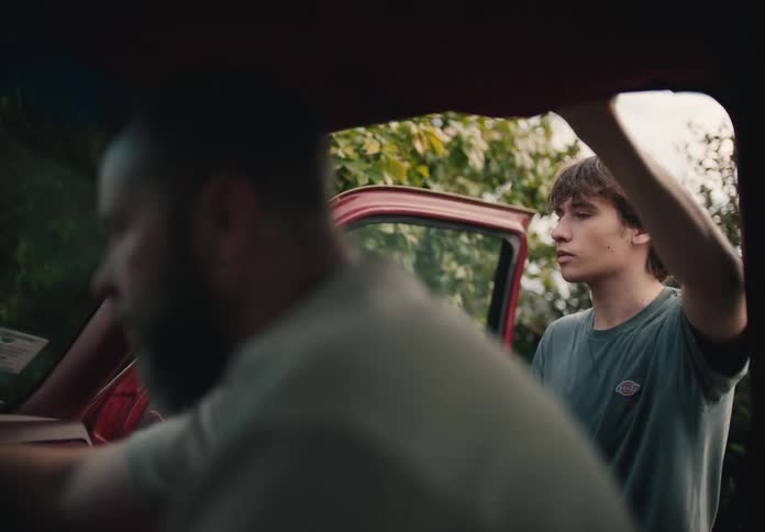A young man leans on the open door of a red truck as his father cleans the interior