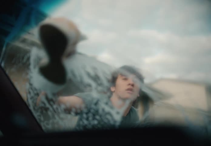A young man washes a car's windshield with a soapy sponge, seen from the driver's seat perspective