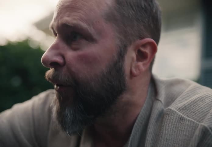A close-up profile of a worried, bearded middle-aged man talking to someone off-screen outdoors
