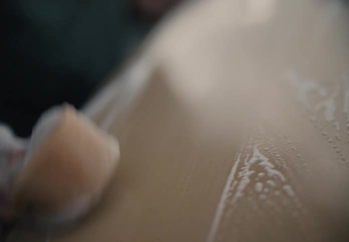 A person scrubs the hood of a car with a soapy sponge in a detailed, slow-motion close-up