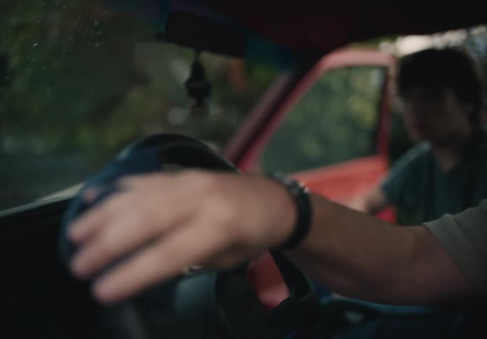 A man meticulously wipes down the steering wheel of a vintage truck as a younger man waits in the doorway