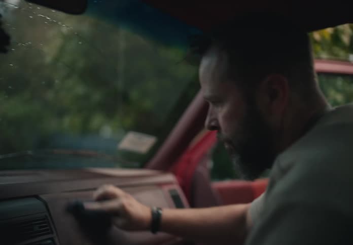 A bearded man meticulously cleans the dusty dashboard of his vintage red truck on an overcast day