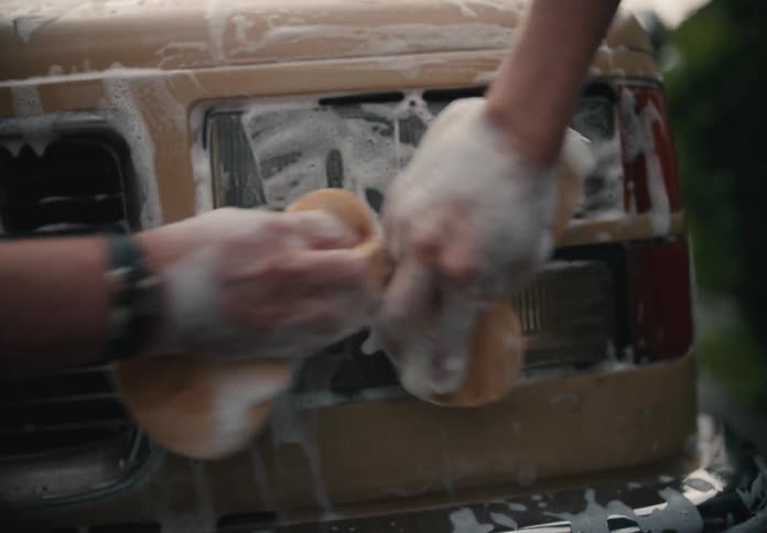 Close-up shot of two people washing the headlights and grille of a vintage tan truck with sponges and soap