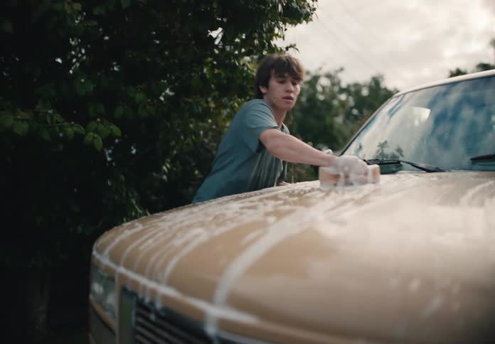 A teenage boy diligently scrubs the hood of a tan truck in a driveway before playfully throwing the soapy sponge