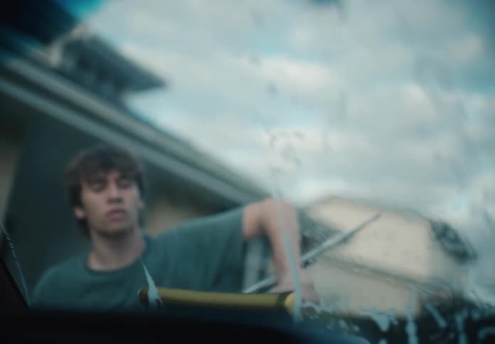 A young man washes a soapy car windshield with a squeegee in slow motion as seen from inside the vehicle