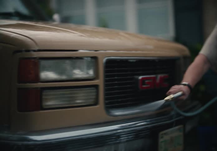 A man sprays down his tan GMC truck with a water hose in his driveway in slow motion